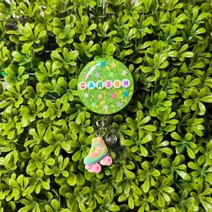 以下が含まれることがあります： カラフルな文字で「CARSON」と書かれた、緑色のグリッターバッジリール。パステルカラーのローラースケートのチャームが下にぶら下がっています。リールは緑の葉の背景に配置されています。