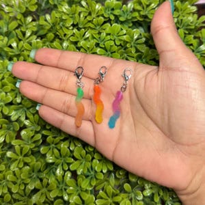 May include: Three colorful gummy worm charms with silver-tone clasps. Each charm features a gradient of colors, including green, orange, yellow, pink, and blue. The charms are held in a hand against a backdrop of green foliage.
