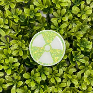 Pode incluir: Um adesivo redondo branco e verde com glitter com um símbolo de radiação. O adesivo é redondo e tem um fundo branco com uma sobreposição de glitter verde.