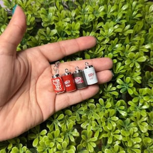 May include: Miniature soda can charms in red, white, and black, including Coca-Cola and Dr. Pepper designs. The charms are attached to a silver chain. The charms are held in a hand against a background of green foliage.