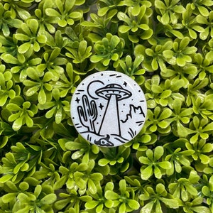 May include: A white and black enamel pin with a cartoon illustration of a UFO hovering over a desert landscape with a cactus. The UFO has a beam of light shining down from it.