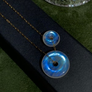 May include: A gold chain necklace with two iridescent moonstone donut pendants. The larger pendant is about 3.8 cm in diameter and the smaller pendant is about 2.5 cm in diameter.