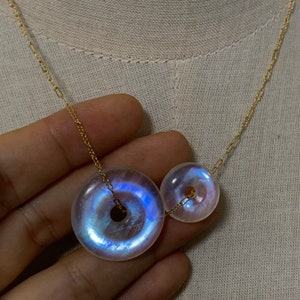 May include: A gold chain necklace with two iridescent moonstone donut pendants. The larger pendant is on the left and the smaller pendant is on the right.