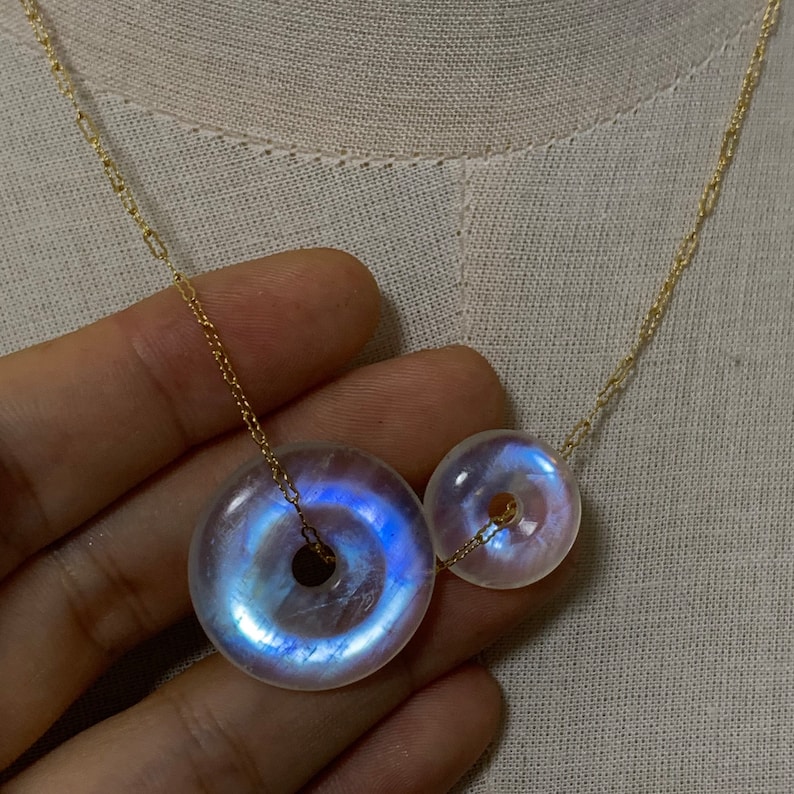 May include: A gold chain necklace with two iridescent moonstone donut pendants. The larger pendant is on the left and the smaller pendant is on the right.