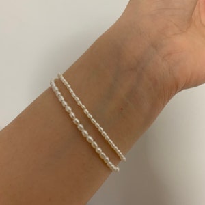 Peut inclure: Deux bracelets en perles sur le poignet d'une personne. Les bracelets sont faits de petites perles blanches et sont superposés l'un sur l'autre.