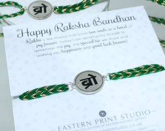 May include: Three green and gold braided bracelets with silver charms that say "bro" in Hindi script. The bracelets are on a white card with the text "Happy Raksha Bandhan" and a message about the meaning of the Rakhi. The card also includes the text "Eastern Print Studio" and a website address.