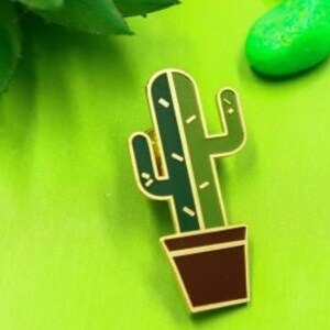 Puede incluir: Un pin de cactus de esmalte verde y marrón con una maceta. El cactus está en una maceta marrón con un borde dorado.