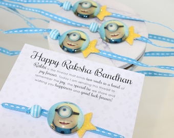 May include: Three blue and yellow Minion-themed Rakhi bracelets with white and blue beads. The bracelets are tied with blue ribbon and have a yellow star charm. The card reads "Happy Raksha Bandhan Rakhi is the thread that binds two souls in a bond of joy forever. Today I am sending this thread to remember the joy, the special tie we share and wishing you happiness and good luck forever!"