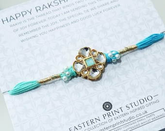 May include: A turquoise Rakhi bracelet with a gold-toned floral centerpiece, adorned with clear and turquoise stones. The bracelet has turquoise thread ends and is presented on a card with text.