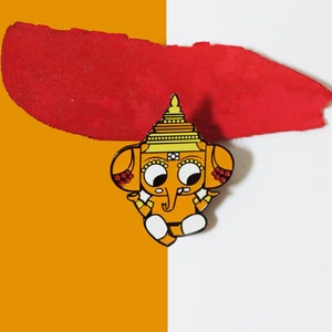 May include: An enamel pin featuring a cartoon-style elephant with a yellow and orange body, a red and yellow crown, and large eyes. The pin is a fun and whimsical way to show your love for elephants.