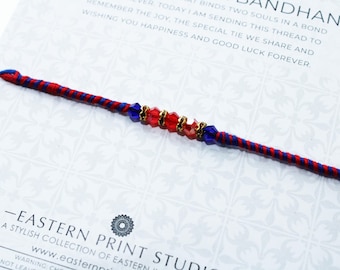 May include: A red and blue thread bracelet with decorative red and blue beads. The bracelet is on a white card with text that reads "BANDHAN" and "EASTERN PRINT STUDIO".