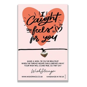 May include: A pink heart with the text "I Caught the Feels for You" on a white background. A black string bracelet with a silver heart charm is attached to the card. The card also includes the text "Make a wish, tie on the bracelet when the thread wears thin & breaks away your wish will come true, so they say." and "Wish Strings. www.wishstrings.co.uk Handmade in the UK."