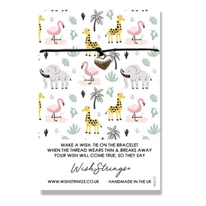 Op de afbeelding: Een zwart koordarmband met een zilveren hartbedel. De armband heeft een herhalend patroon van giraffen, olifanten, flamingo's, palmbomen en cactussen. De tekst "Make a wish, tie on the bracelet when the thread wears thin & breaks away your wish will come true, so they say" staat op de kaart gedrukt. De tekst "Wish Strings" staat in een gestileerd lettertype gedrukt. De tekst "www.wishstrings.co.uk Handmade in the UK W$272" staat onderaan de kaart gedrukt.