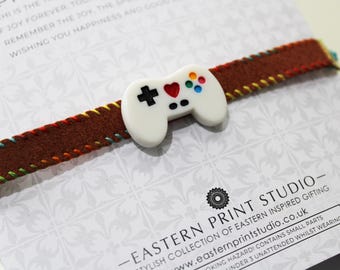 May include: A brown bracelet with a white controller charm. The bracelet has colorful stitching along the edges. The controller charm has a heart, cross, and colorful buttons. The text on the card reads "EASTERN PRINT STUDIO".