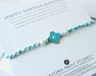 May include: A turquoise blue and white braided string bracelet with a gold flower charm and white pearl beads. The bracelet is tied with a knot and has the text "HAPPY RAKSHA BANDHAN" on the card behind it.