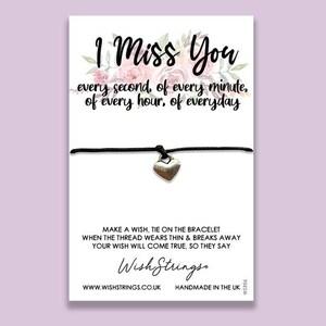 May include: A white card with the text "I Miss You" and a floral design. Below is a black string bracelet with a silver heart charm. The card includes text about making a wish and the Wish Strings logo.