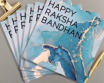 May include: A stack of blue and gold cards with the text "Happy Raksha Bandhan" printed on the top card.