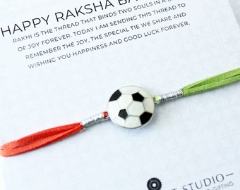May include: A green, red, and silver Raksha Bandhan bracelet with a soccer ball charm. The bracelet has the text "HAPPY RAKSHA BANDHAN" and a message about the bond between two souls.