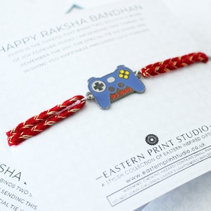 Puede incluir: Un brazalete de cuerda rojo y dorado con un dije de mando de juego azul y plateado. El dije tiene las palabras "PS BRO" en él. El brazalete está sobre un fondo blanco con el texto "HAPPY RAKSHA BANDHAN" y "EASTERN PRINT STUDIO" en él.