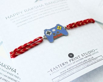May include: A red and gold string bracelet with a blue and silver game controller charm. The charm has the words "PS BRO" on it. The bracelet is on a white background with the text "HAPPY RAKSHA BANDHAN" and "EASTERN PRINT STUDIO" on it.