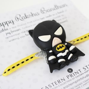 May include: A yellow and black patterned ribbon with a black and white Batman character charm. The charm has a yellow Batman logo on the chest. The ribbon is tied around a white card with the text "Happy Raksha Bandhan" and a message about joy and wishing someone well. The card also has the text "Eastern Print Studio" and a website address.