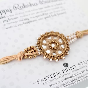May include: A gold and white Rakhi bracelet with a circular design and a tassel. The bracelet is on a white card with the text "Happy Raksha Bandhan" and "Eastern Print Studio".