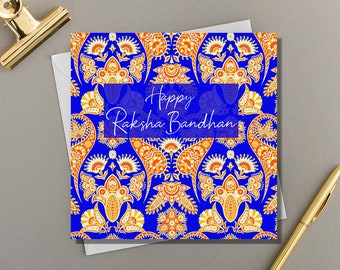 May include: Square greeting card with a blue background and an orange and white floral pattern. The text "Happy Raksha Bandhan" is displayed in white script within a blue rectangle. A gold pen and paper clip are visible.