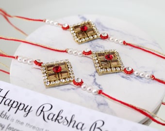 May include: Three red and gold Rakhi bracelets with white pearl beads and a red stone in the center of each square charm. The bracelets are on a white surface with a card that says "Happy Raksha Bandhan".