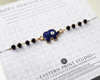 May include: A gold-toned bracelet featuring a blue elephant charm with an eye design. The bracelet also has black beads. The bracelet is displayed on a patterned card with text.