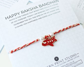 May include: Red and gold braided string bracelet with a red airplane charm and white beads. The bracelet is tied around a card with the text "Happy Raksha Bandhan" and a message about the special bond between siblings.