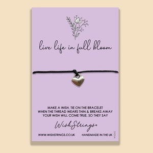 May include: A purple card with the text "live life in full bloom" and a black string bracelet with a silver heart charm. The card also includes the text "Make a wish, tie on the bracelet when the thread wears thin & breaks away your wish will come true, so they say." and the logo "Wish Strings."