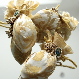 Op de afbeelding: Close-up van verschillende kleine, decoratieve stoffen zakjes. De zakjes zijn wit met een gouden bloemenpatroon en zijn vastgebonden met gouden touw. Elk zakje is versierd met een cluster van gouden kralen en een klein juweel met een pauwenveerontwerp.