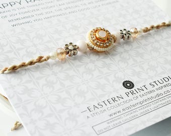 May include: A Rakhi bracelet with a central gold-colored, circular charm adorned with small white beads and a pink stone. The bracelet features decorative silver-colored flower accents and a twisted, beige cord.