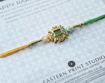 May include: A gold-colored Rakhi bracelet with a central square green stone and surrounding clear stones. The bracelet has a yellow and green thread design with small white pearls. The background has text that reads "HAPPY RAKHI".