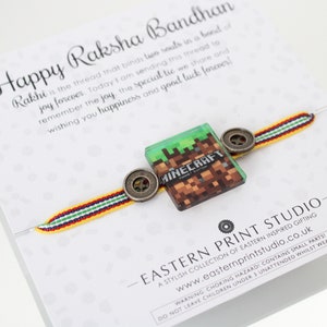 May include: A yellow, red, and green braided bracelet with a Minecraft logo charm and two metal buttons. The bracelet is tied around a card with the text "Happy Raksha Bandhan" and a message about the special bond between siblings.