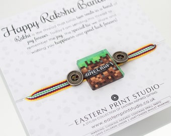 May include: A yellow, red, and green braided bracelet with a Minecraft logo charm and two metal buttons. The bracelet is tied around a card with the text "Happy Raksha Bandhan" and a message about the special bond between siblings.