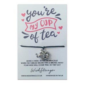 Op de afbeelding: Een zwart koordarmband met een zilveren theepotbedel. De kaart die aan het armband is bevestigd, luidt "you're my cup of tea". Doe een wens, bind het armband om. Wanneer de draad dun wordt en breekt, zal je wens uitkomen, zeggen ze. Wish Strings. www.wishstrings.co.uk Handgemaakt in het Verenigd Koninkrijk