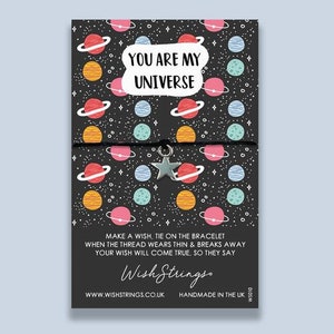 May include: A black and white card with a colorful space-themed pattern. The card has a silver star charm attached to a black string. The text on the card reads "You are my universe. Make a wish, tie on the bracelet when the thread wears thin & breaks away your wish will come true, so they say. Wish Strings. www.wishstrings.co.uk Handmade in the UK WS010"