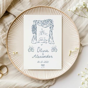 Puede incluir: Una tarjeta de invitaci&oacute;n de boda con un dise&ntilde;o de arco floral azul, con los nombres Olivia & Alexander y la fecha 06-12-2025. La tarjeta est&aacute; sobre un plato beige, con anillos dorados y flores blancas.
