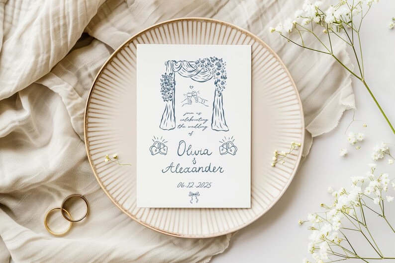 Puede incluir: Una tarjeta de invitaci&oacute;n de boda con un dise&ntilde;o de arco floral azul, con los nombres Olivia & Alexander y la fecha 06-12-2025. La tarjeta est&aacute; sobre un plato beige, con anillos dorados y flores blancas.