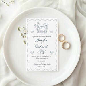 Puede incluir: Una invitaci&oacute;n de boda con una ilustraci&oacute;n de coche y los nombres "Amalia & Richard". Dos anillos de boda dorados descansan junto a la invitaci&oacute;n en un plato blanco, con flores blancas y tela en el fondo.