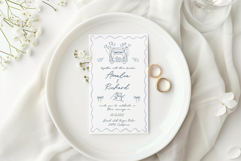 Puede incluir: Una invitaci&oacute;n de boda con una ilustraci&oacute;n de coche y los nombres "Amalia & Richard". Dos anillos de boda dorados descansan junto a la invitaci&oacute;n en un plato blanco, con flores blancas y tela en el fondo.