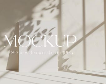 May include: A white mockup card with the word "MOCKUP" in a white sans-serif font. The text "PSD file with smart object + JPEG" is below the word "MOCKUP". The card is sitting on a white surface with shadows from a window.
