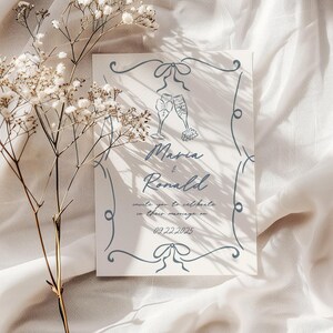 Puede incluir: Una invitaci&oacute;n de boda con borde y texto azules. La invitaci&oacute;n presenta dos copas de champ&aacute;n y los nombres "Maria & Ronald". Tambi&eacute;n se incluye la fecha "02.22.2025". Flores secas en primer plano.