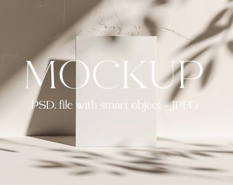 May include: A white mockup card with the word "MOCKUP" in white text and the text "PSD file with smart object + JPEG" in white text. The card is sitting on a light beige surface with shadows from a window.