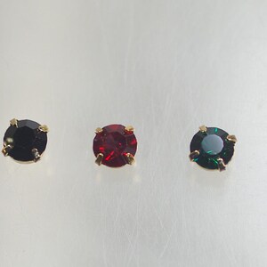 Könnte beinhalten: Fünf goldfarbene Ohrstecker mit runden, facettierten Edelsteinen. Die Edelsteine sind irisierend, schwarz, rot, dunkelgrün und bernsteinfarben. Die Ohrringe sind vor einem hellen Hintergrund.