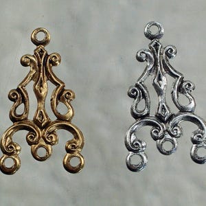 May include: Two ornate metal chandelier components. One is gold-toned, the other silver-toned. Each has a decorative scroll design with a loop at the top and three loops at the bottom. These are likely for jewelry making or craft projects.