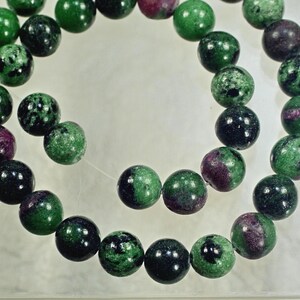 Puede incluir: Un collar de cuentas redondas de piedras preciosas. Las cuentas son una mezcla de verde oscuro y rojo púrpura intenso, con un patrón moteado. Las cuentas están ensartadas en un cordón fino y transparente, de aproximadamente 8 mm de diámetro.