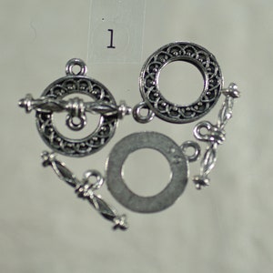 Puede incluir: Un conjunto de cierres de palanca plateados para la creación de joyas. Los cierres presentan intrincados diseños de filigrana e incluyen componentes circulares y cierres de barra. El número "1" es visible en la esquina superior izquierda.