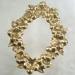 Op de afbeelding: Een goudkleurige, ovaalvormige hanger of bedel, versierd met gedetailleerde bloemen- en bladerontwerpen. Het metalen stuk heeft een lus aan de bovenkant om op te hangen en is waarschijnlijk gemaakt van messing of een vergelijkbaar materiaal. De bloemen en bladeren zijn ingewikkeld gebeeldhouwd.